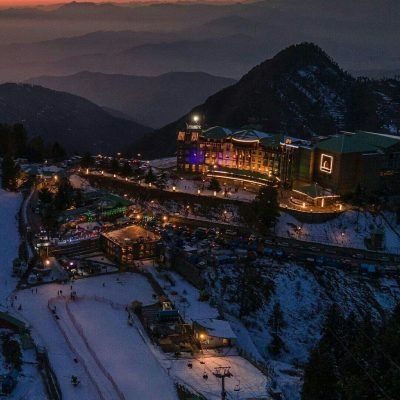 Snowy ski slope at sunset with lit resort buildings and mountain ridges in background